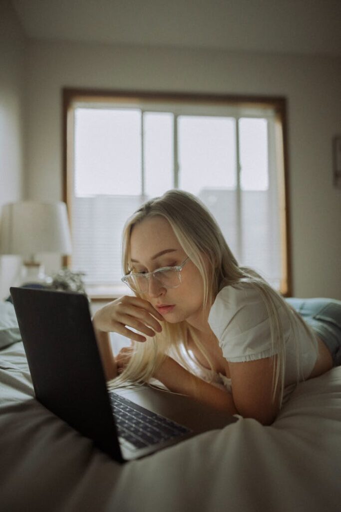 Young woman in eyeglasses using laptop while lying on bed in a softly lit room.