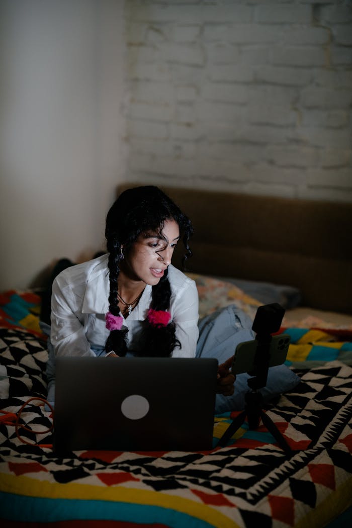 Young woman with braided hair lying on bed using laptop and smartphone with tripod.