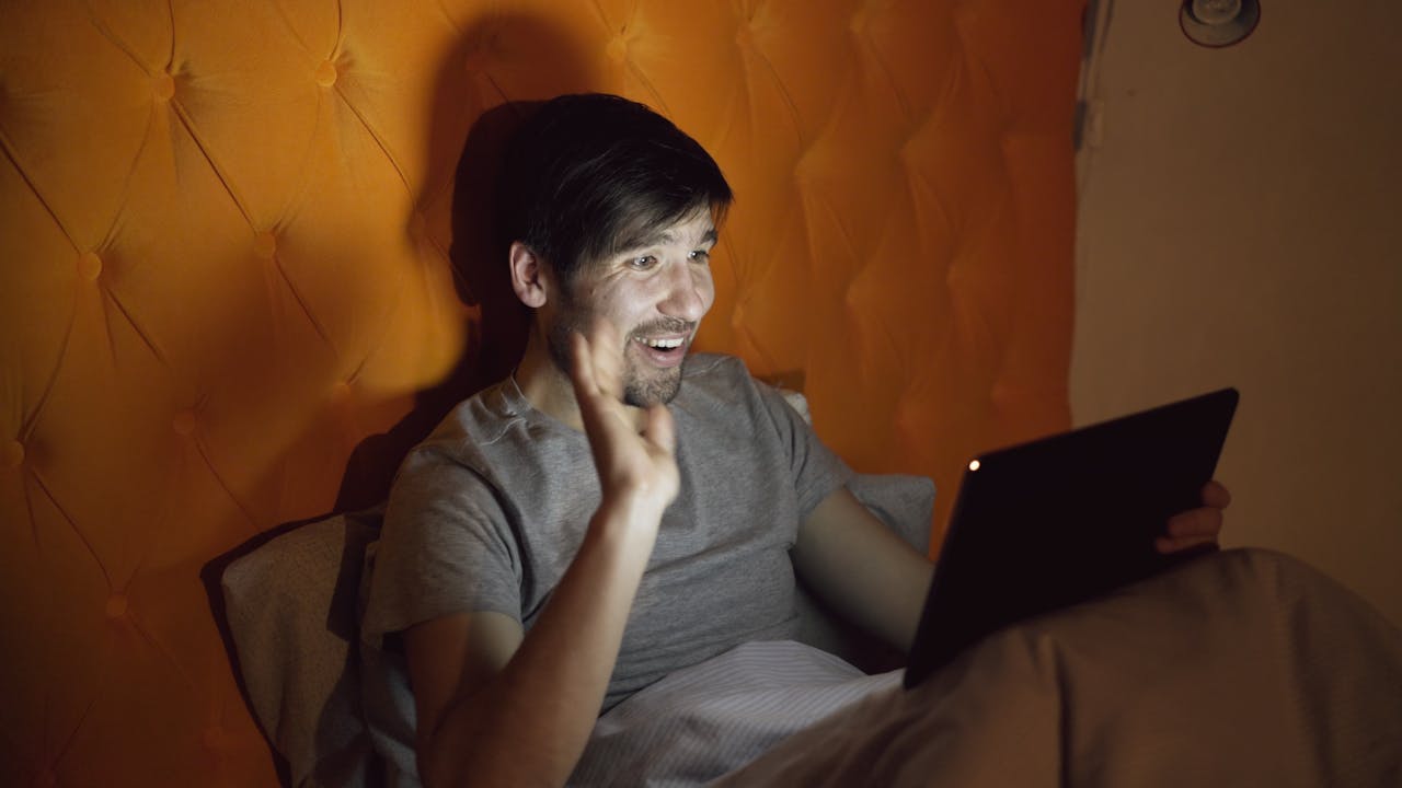 A man in bed using a tablet at night, actively video calling.