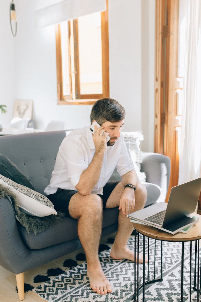 Adult man sitting on a sofa, making a phone call while working on a laptop indoors.