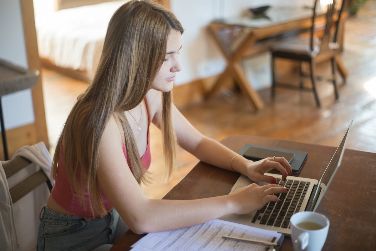 Woman with long hair typing on laptop indoors, coffee nearby, cozy home environment.
